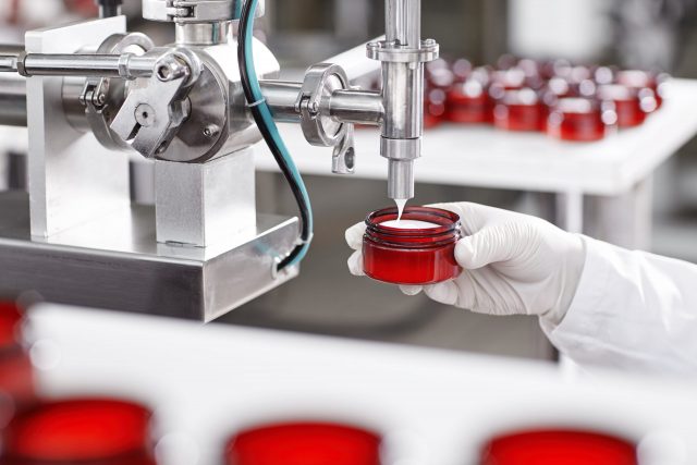 Portrait of big factory process of creating lotion for skincare. Researcher holding container under big metal tube pouring beauty cream isolated over working process at factory and many containers